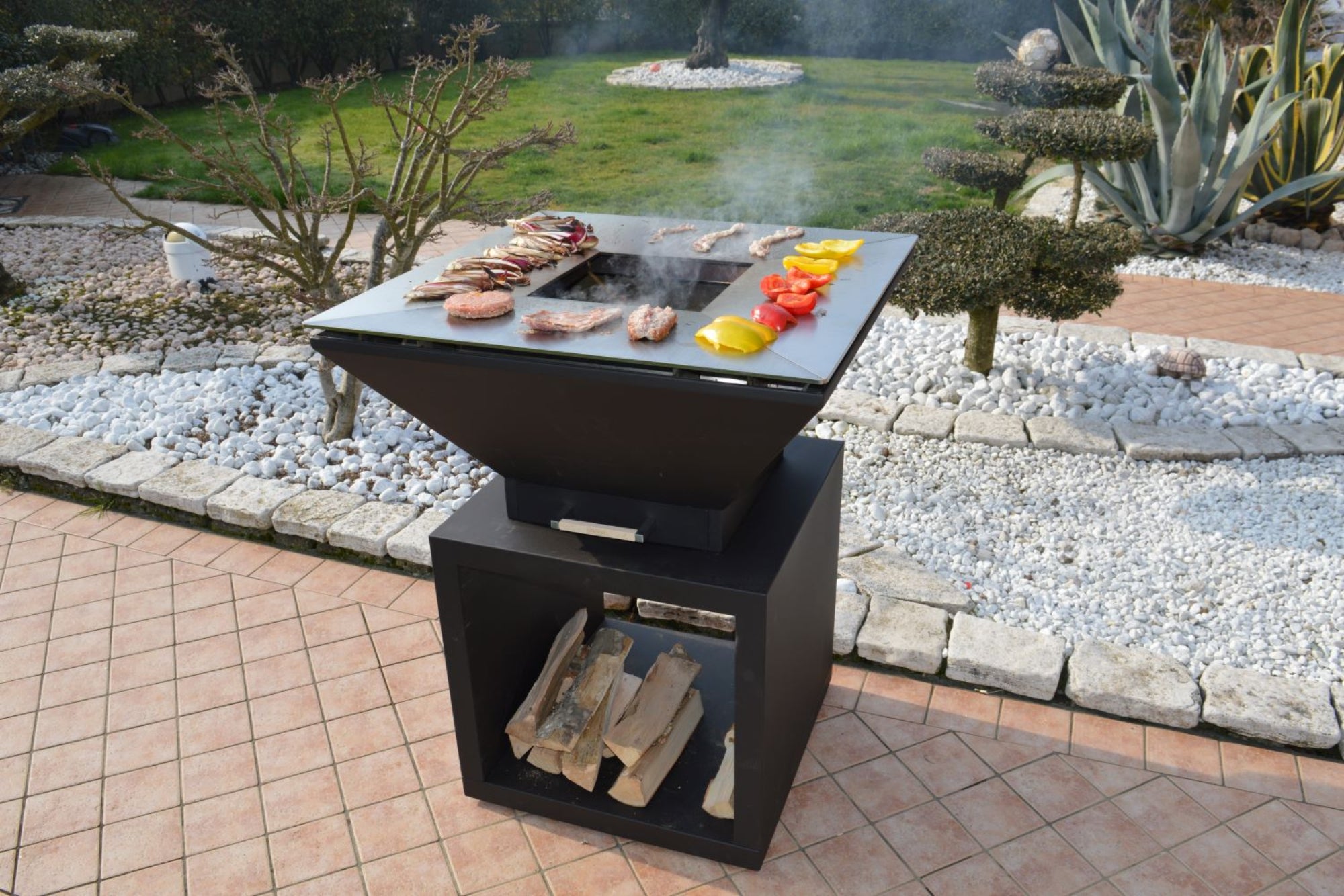 A charcoal grey BBQ tower with wood storage, featuring an open cover, cooking grill, and grilled plate with food items on top, set against an outdoor backdrop.